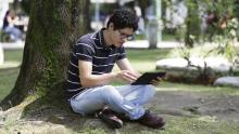 Un joven consulta una tableta digital sentado bajo un árbol